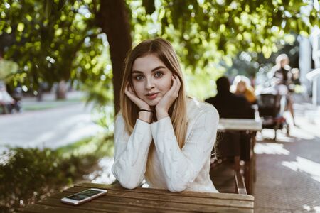 girl with big beautiful eyes propping her hands on her cheeks is sitting in a street cafeの写真素材