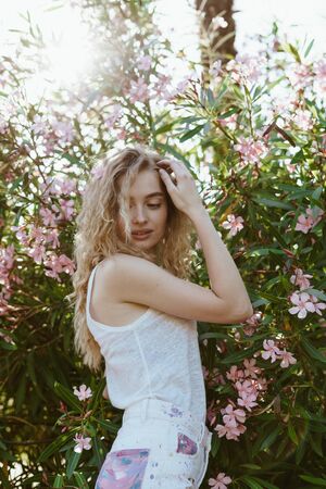 blonde straightens her curly hair with her hand and looks embarrassed down standing by a bush with blooming oleander flowersの写真素材