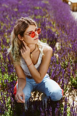 blonde with sportswear squatted among sage flowers and adjusts the earphone in her ear looking into the distance through pink glassesの写真素材