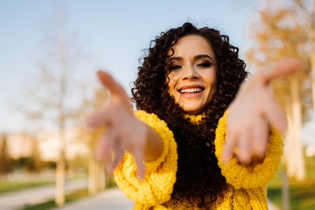 Curly brunette with a charming smile pulls her hands into the camera in a bright yellow sweater against the background of autumn trees. High quality photoの写真素材