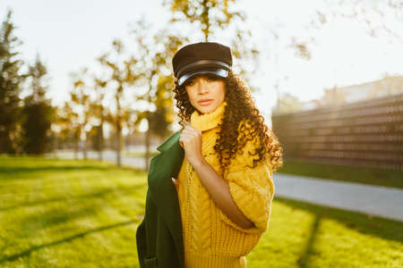 In the autumn park, a girl in a leather cap and a bright yellow sweater slung over her shoulder a green jacket with her hair with dark curls scattered over her shoulders. High quality photoの写真素材