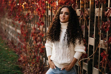 A beautiful brunette in a white sweater and blue jeans holds her hands in her front pockets against the wall of entwined red ivy. High quality photoの写真素材