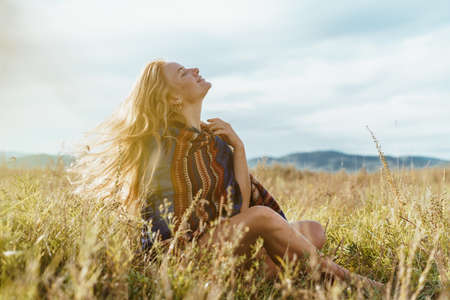 Bright red girl enjoys the sun, sits in a field among the grasses against the backdrop of mountains...
