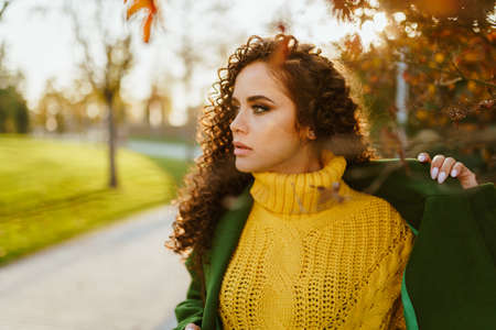 A strong look at the face of the curly brunette in a bright yellow sweater and green coat against the backdrop of the autumn park. High quality photoの写真素材