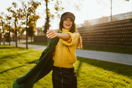 Stretching forward one hand and the other covering the coat thrown over her shoulders, the girl happily calls someone in the autumn park dressed in bright stylish clothes. High quality photoの写真素材