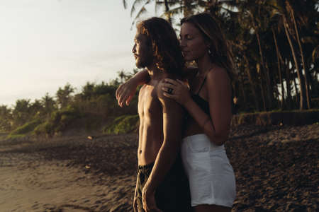 On the coastal black sand of Bali there is a beautiful couple in love. Woman of European appearance hugs from the back of a naked torso of a man of East Asian appearance. High quality photoの写真素材