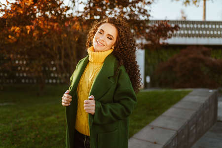 Curly brunette with a lovely smile poses in the autumn park holding hands by the lapels of her coat....