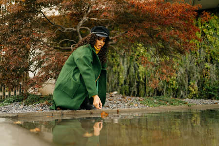 At the backwater in the park crouched a girl in an autumn coat and a cap of curly dark hair scattered over the shoulders she reaches out with her hand to the water. High quality photoの写真素材
