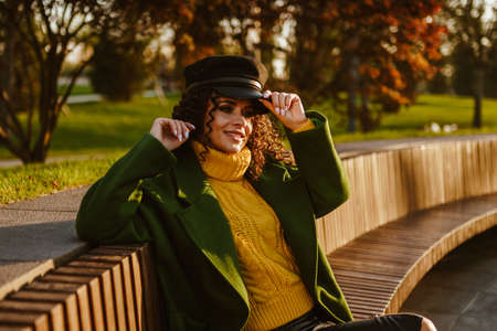 The beauty stylish with a confident white-toothed smile holds the hand visor of his cap sitting on a bench in the park in bright autumn clothes. High quality photoの写真素材