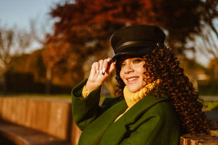 The beauty in the autumn park in a coat holds her hand behind the visor of the cap and smiles. High...