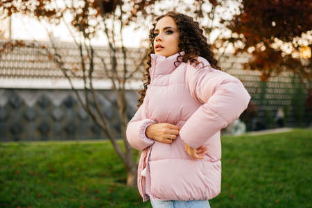 Thoughtfully looking into the distance, a girl in a down jacket stretches her hand into her pocket against a backdrop of park grass. High quality photoの写真素材