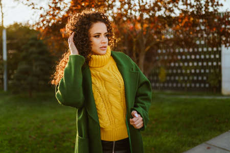 Luxury beauty in a bright yellow sweater and dark green coat stands on the green grass of the park. High quality photoの写真素材