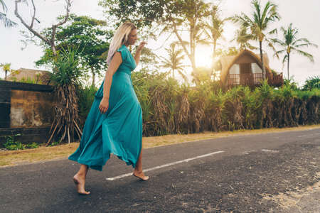 A girl of size in a long blue dress runs barefoot on the track against a backdrop of palm trees and villas. High quality photoの写真素材