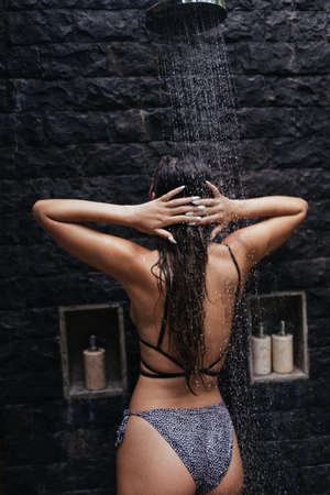 photo from the back close-up. A girl in a swimsuit takes a shower at a dark stone wall. High quality photoの写真素材