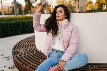 A young woman tucked under herself one leg sits on a bench in the park and plays with her hair curls. High quality photoの写真素材
