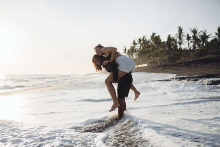 a man walks along the waters edge in the ocean and carries a woman on her shoulders against the backdrop of coastal palm trees. High quality photoの写真素材
