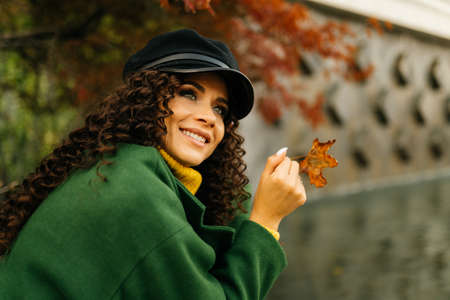 Beautiful curly brunette in a cap with a visor and autumn green coat with a charming smile turned...