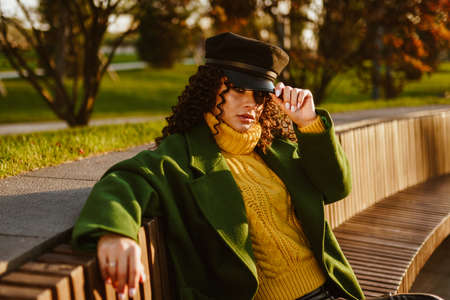on a long wooden bench in the park sits a girl in autumn clothing holding on to the visor of a cap...
