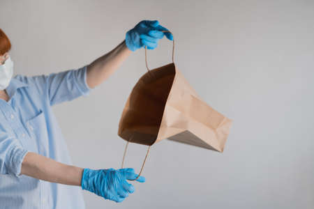 A woman in a medical mask looks into an empty paper bag on a white background. Woman in blue latex...