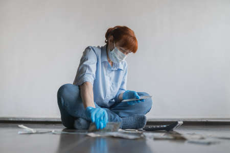 An adult woman in a medical mask on her face sits on the floor and counts money on a calculator, on...