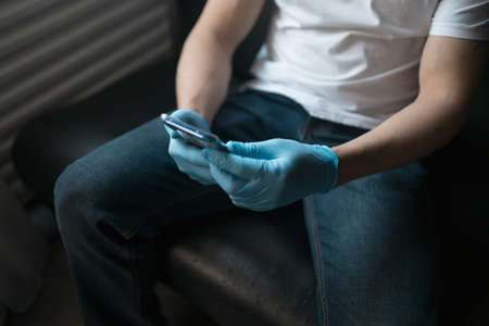 A man uses a smartphone while sitting on the couch. Blue latex gloves and white jersey. High quality photoの写真素材