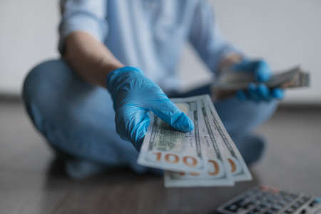 A hand in a blue medical glove holds a wad of dollars and points to the camera. Blurred figure in the background. High quality photoの写真素材
