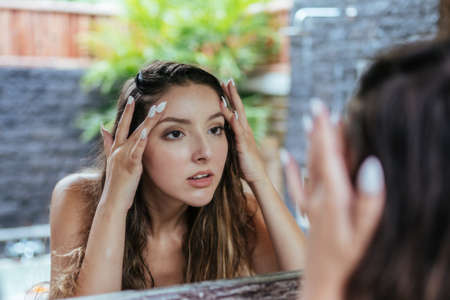 In the bathroom at the mirror the girl touches her face with her fingers looking at her skin in search of pimples or wrinkles. High quality photoの写真素材