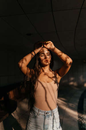 A young girl in a beige T-shirt and white jeans touches her head with her hands. Shot in the setting sun. Evening walk in the city autumn park. Portrait of a girl to the waist. High quality photoの写真素材