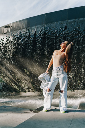 A girl with dark long hair in glasses, a beige T-shirt in white jeans and a white jacket in her hands. A young girl poses against the background of a fountain in the park. The girl touches her head with her right hand and lifted her head up to the left. Summer walk in the park in the fresh air. High quality photoの写真素材