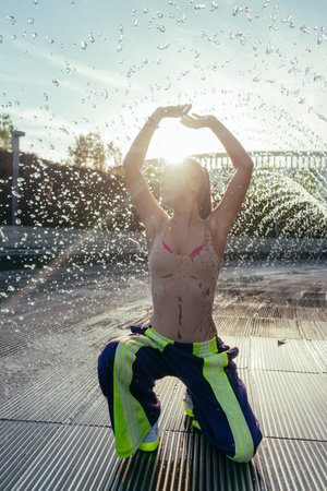 Photo of a young sports girl in a fountain. A girl in a beige T-shirt and purple pants with bright stripes. Shot against the sun. The happy girl raised her hands up and sat down, looking to the left. A beautiful shot in the park with splashes of a fountain in the background. A girl in a wet T-shirt. High quality photoの写真素材