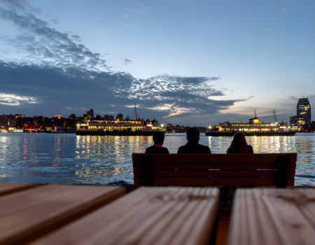 Silhouette of three people on a bench enjoying a tranquil harbor view with an illuminated city skyline and ferries at duskの素材