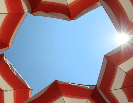 Summer vacation concept with a view of the bright sun and clear blue sky framed by red and white striped beach umbrellas.の素材