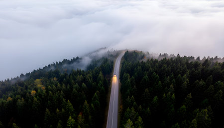 A solitary journey into mystery, aerial view of a car driving on a forest road straight into a thick blanket of fog and clouds.の素材