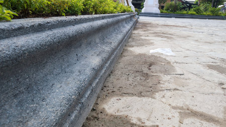 A close-up perspective of a textured gray concrete curb bordering a flower bed, with sandy earth and construction materials on the ground.