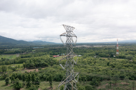 High-voltage power lines and transmission pylon towering over rural landscape with verdant trees. Showcasing electricity distribution, energy infrastructure, utility grid.の写真素材