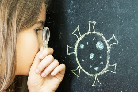 Little girl alone at home showing observing drawing of a coronavirus on a blackboard with a magnifying glassの写真素材