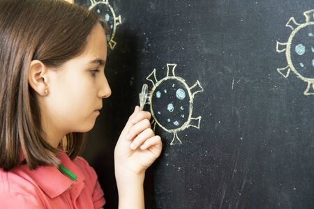 Little girl alone at home showing observing drawing of a coronavirus on a blackboard with a magnifying glassの写真素材