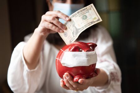 Woman with mask, worried about confinement due to the covid19 pandemic, inserting dollars into a pig-shaped piggy bank also with maskの写真素材