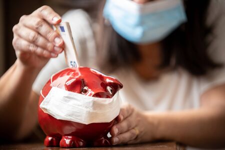 Woman with mask, worried about confinement due to the covid19 pandemic, inserting euros into a pig-shaped piggy bank also with maskの写真素材