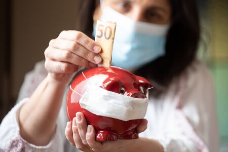 Woman with mask, worried about confinement due to the covid19 pandemic, inserting euros into a pig-shaped piggy bank also with maskの写真素材