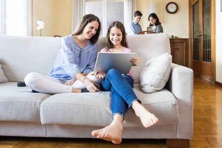 Two sisters with a tablet sitting on the sofa at home with the mother and another brother talking in...