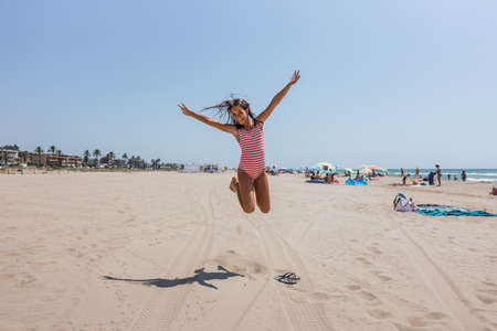 A smiling girl in a pink white striped swimsuit jumping with hands up on the sand in summer and there is a lot of people in the backgroundの写真素材