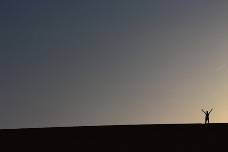 A lonely of adventurer salutes from the top of a grand sand dune while enjoying a beautiful summer sunset in Dakhla Oasis, Egyptの写真素材