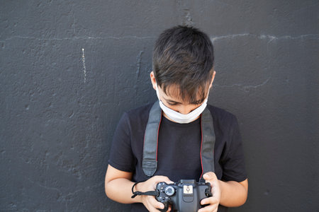 A cute little kid with green eyes wearing a mask is holding a camera and taking pictures. Happy boy dreams of becoming a photographer.の写真素材