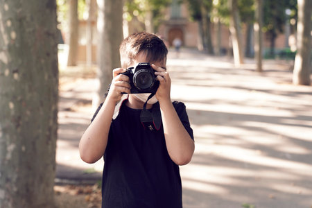 A cute little kid with green eyes wearing a mask is holding a camera and taking pictures. Happy boy dreams of becoming a photographer.の写真素材