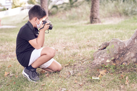 A cute little kid with green eyes wearing a mask is holding a camera and taking pictures. Happy boy dreams of becoming a photographer.の写真素材