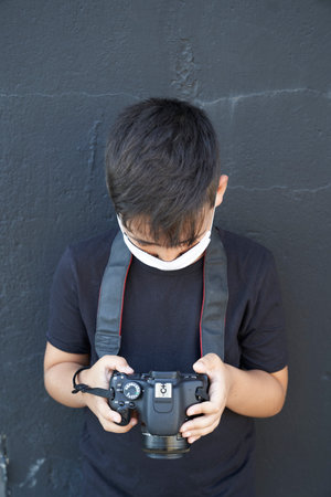 A cute little kid with green eyes wearing a mask is holding a camera and taking pictures. Happy boy dreams of becoming a photographer.の写真素材