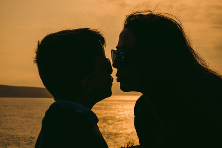 Silhouette of a young mother lovingly kissing her little child, outside isolated in front of a sunset in the sky on a summer dayの写真素材