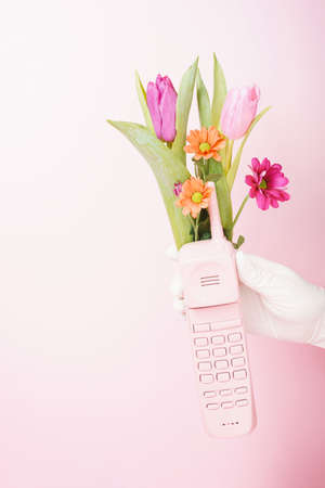 Spring real unique still life composition. Hand with glove holdes a pink telephone and flowers. Advertisement idea. Minimal nature concept.の写真素材