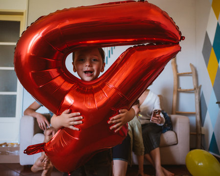 Little boy with big red inflatable number 4 celebrates his birthday at home. In the background is his family sitting on the couch.の写真素材
